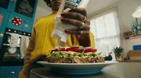 a woman in a yellow sweater is pouring dressing onto a sandwich