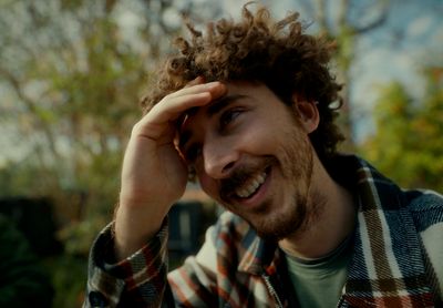 a man with curly hair smiles while holding his hand to his head