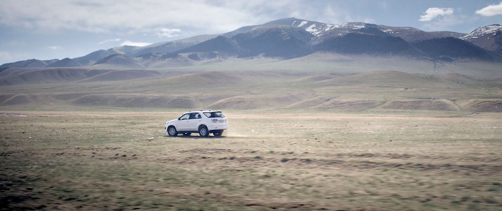 a small white car driving through a field with mountains in the background