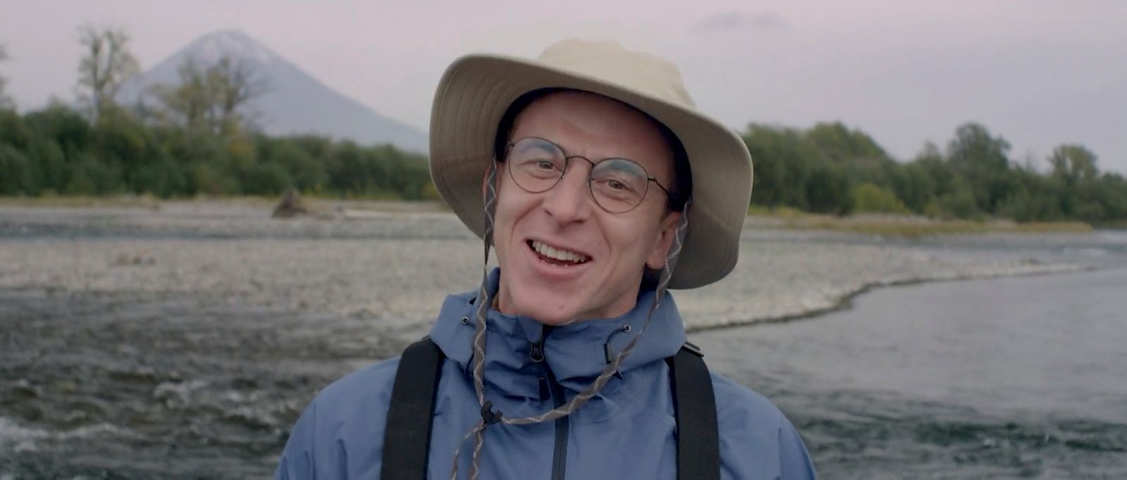 a man wearing a hat and glasses standing in front of a body of water