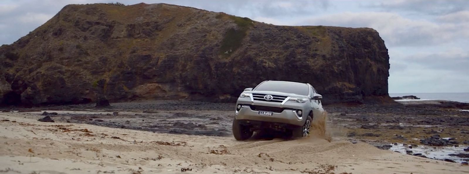 a white truck driving on a sandy beach next to a large rock