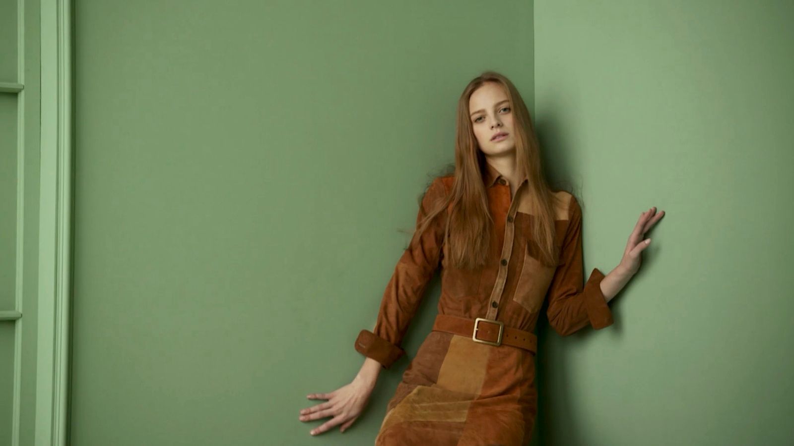 a woman in a brown dress leaning against a green wall