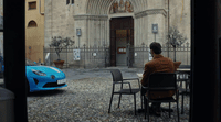 a man sitting at a table in front of a blue sports car