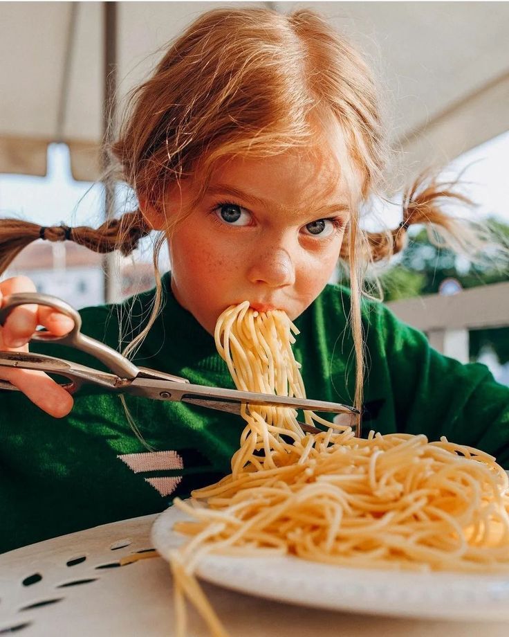 a young girl eating spaghetti with a pair of tongs