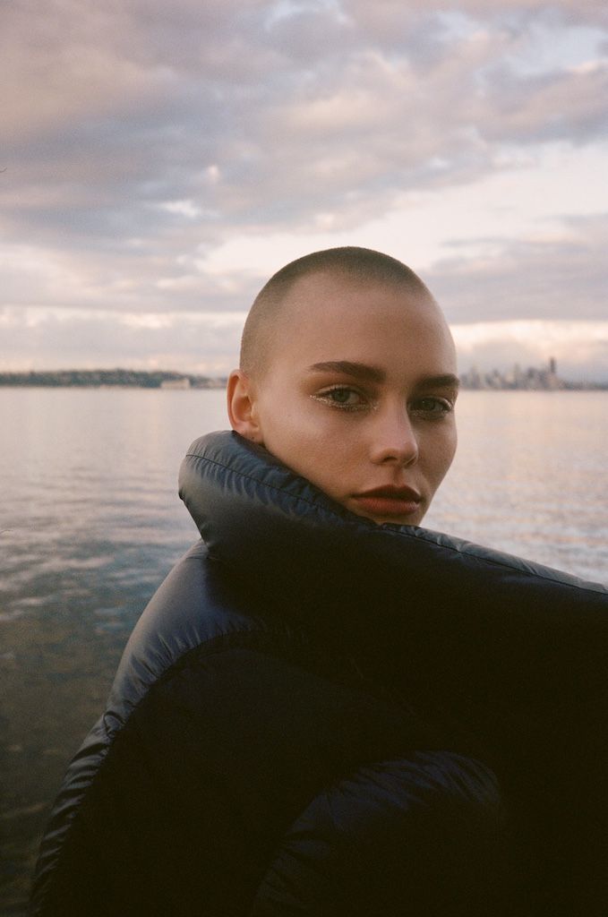 a woman with a shaved head standing in front of a body of water