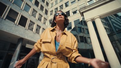 a woman in a yellow coat and sunglasses standing in front of a tall building