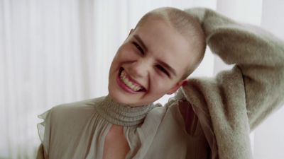 a woman with a shaved head smiles while wearing a sweater