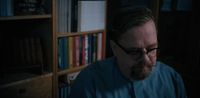 a man with glasses and a beard sitting in front of a book shelf