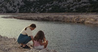 a woman kneeling down next to a little girl near a body of water