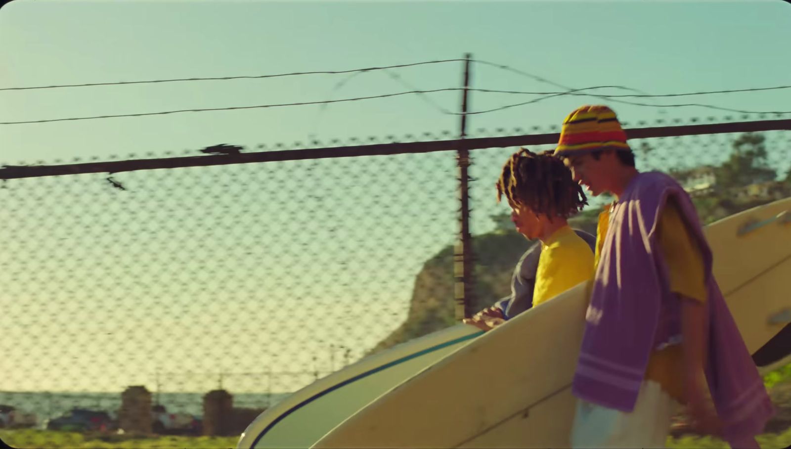 two people carrying surfboards behind a fence