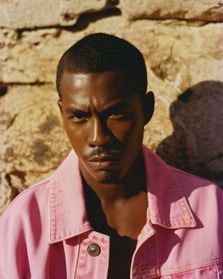 a man in a pink shirt standing in front of a stone wall
