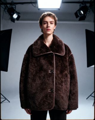 a man standing in a studio wearing a brown coat