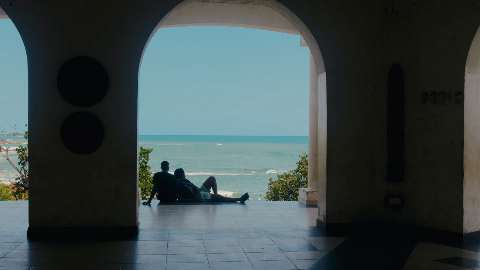 a man and woman sitting on the floor of a building looking out at the ocean