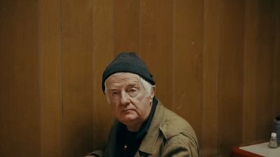 an older man wearing a hat sitting in front of a wooden wall