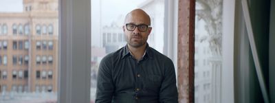 a bald man with glasses standing in front of a window