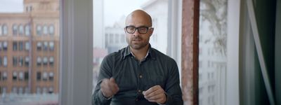 a bald man wearing glasses standing in front of a window