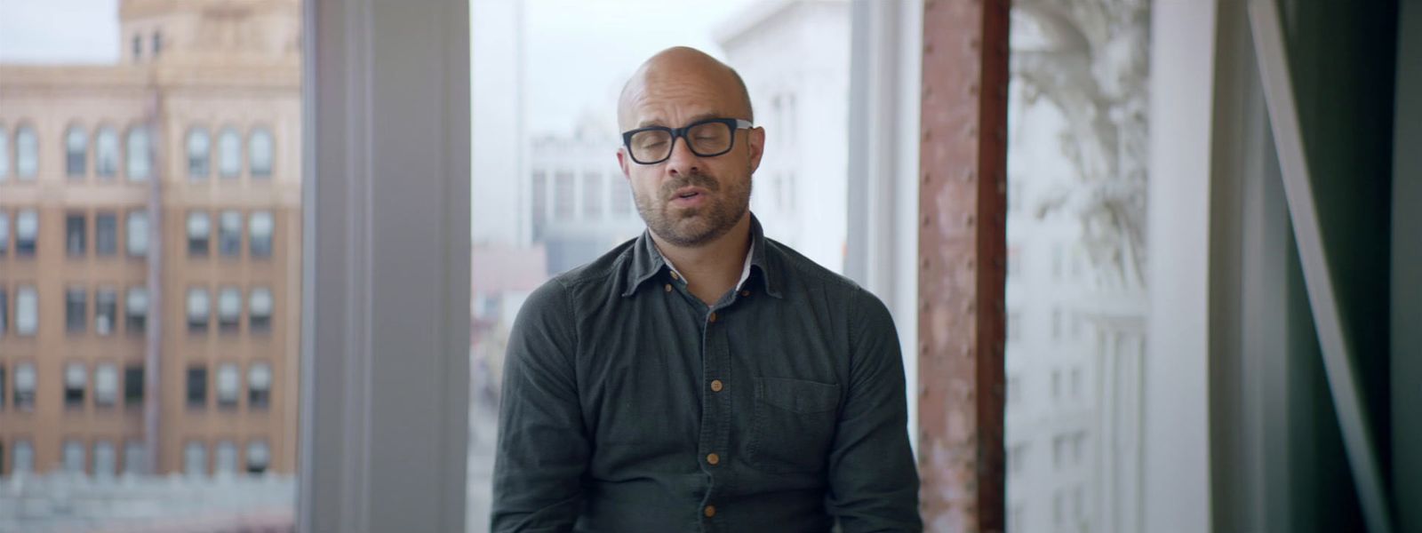 a bald man wearing glasses looking out a window