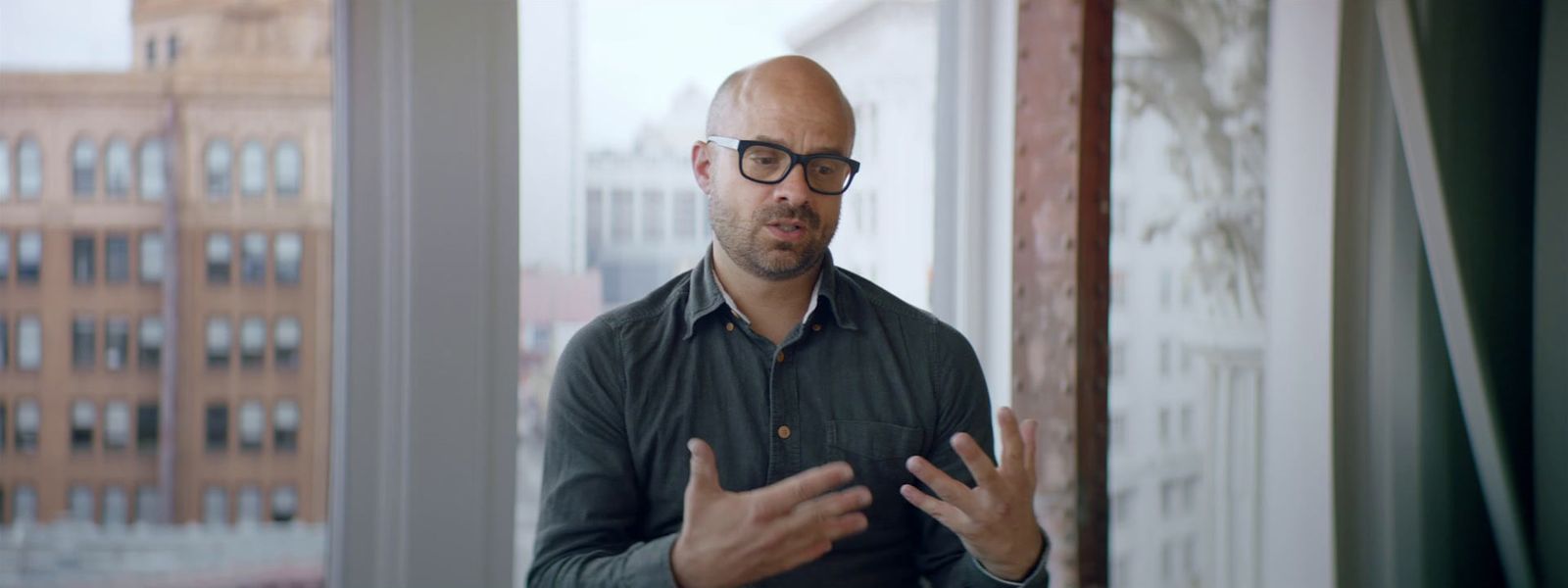 a bald man wearing glasses standing in front of a window