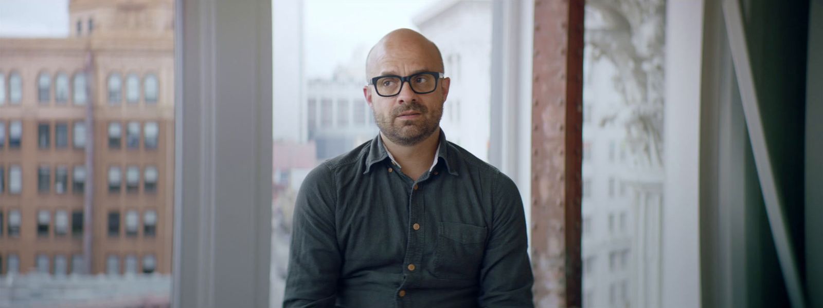 a bald man with glasses standing in front of a window