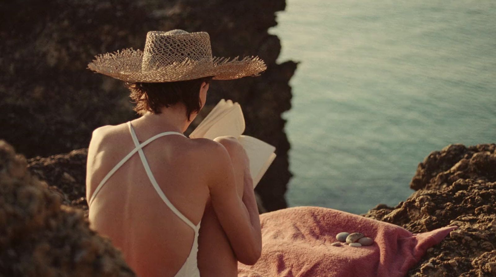 a woman in a straw hat sitting on a rock by the ocean