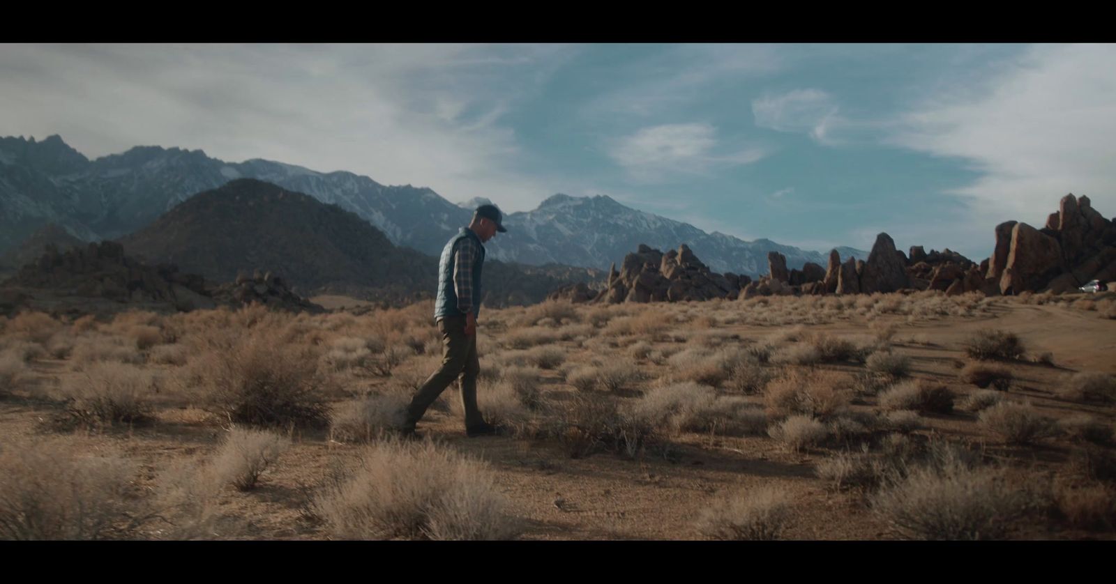 a man walking through a desert with mountains in the background