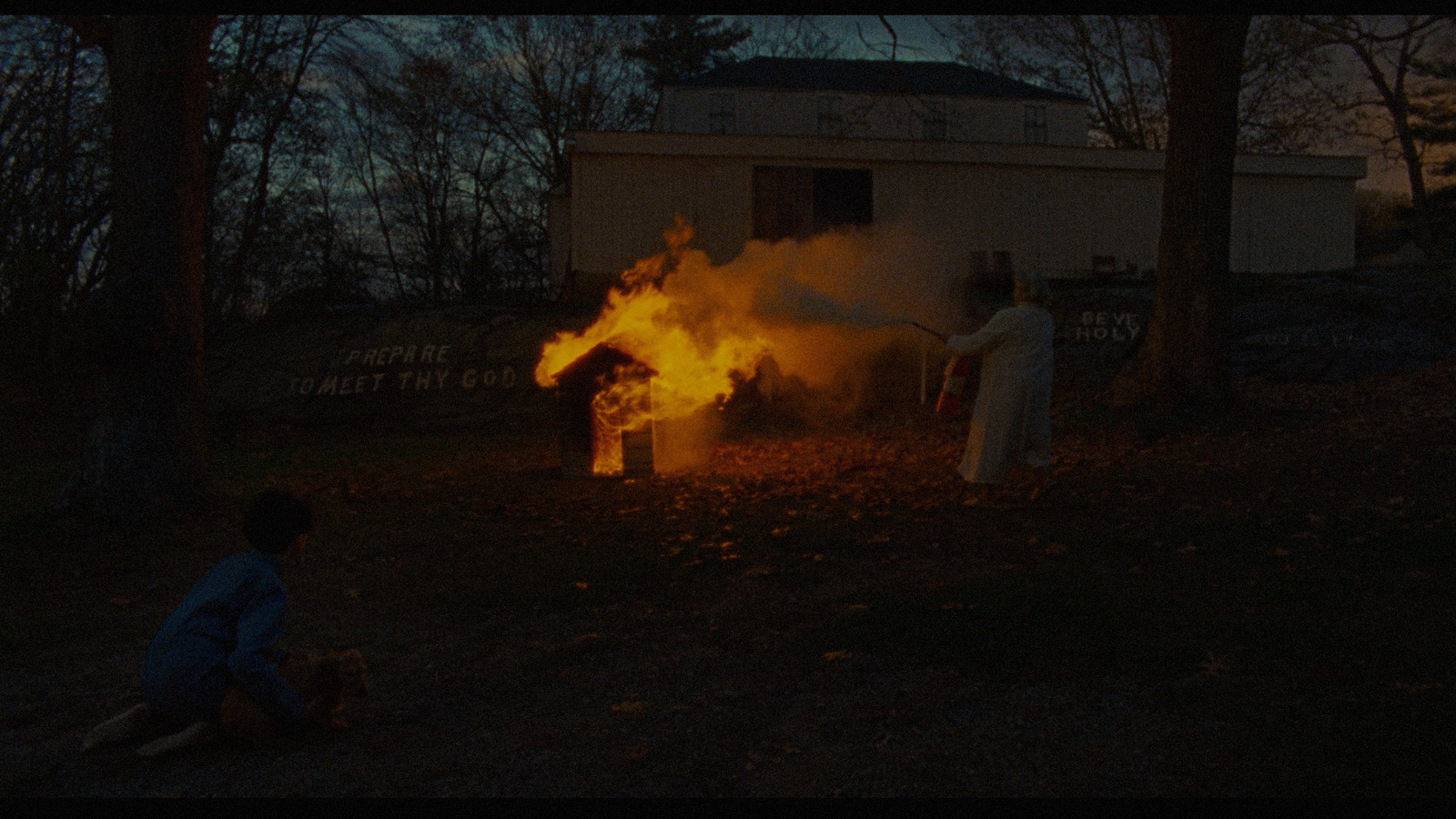 two people sitting in front of a fire with a house in the background