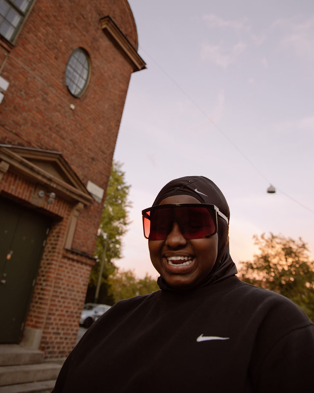 a man wearing sunglasses and a hoodie standing in front of a brick building