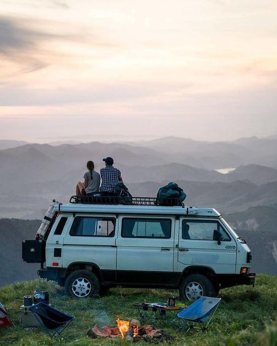 two people sitting on top of a van on top of a hill