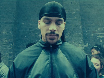 a man in a black hat and jacket standing in front of a brick wall