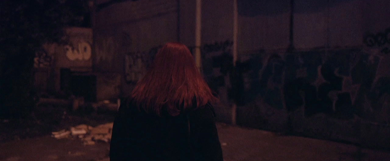 a woman with red hair walking down a street at night