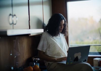 a woman sitting in a kitchen using a laptop computer