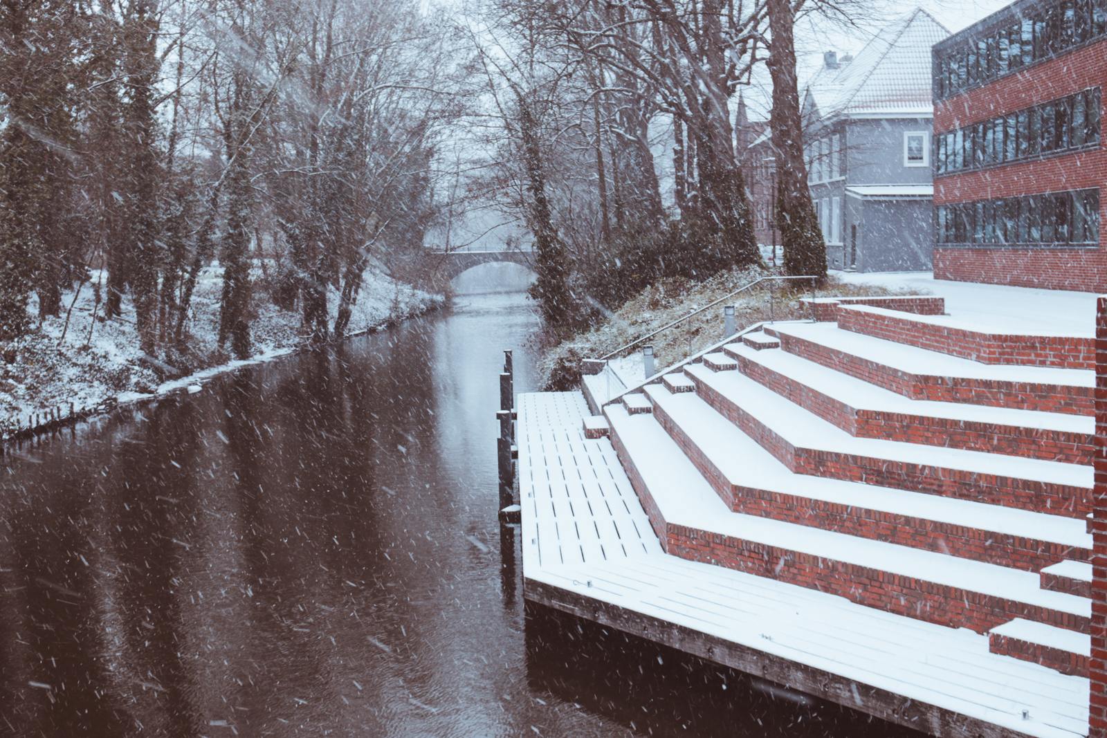 a snowy scene of a river and a red brick building