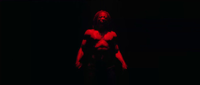 a man with long hair and no shirt standing in the dark