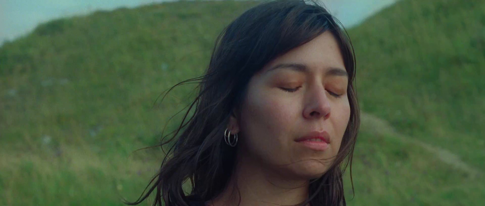 a woman with her eyes closed standing in front of a grassy hill