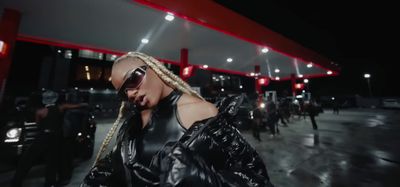 a woman in a black leather outfit and sunglasses standing in front of a gas station