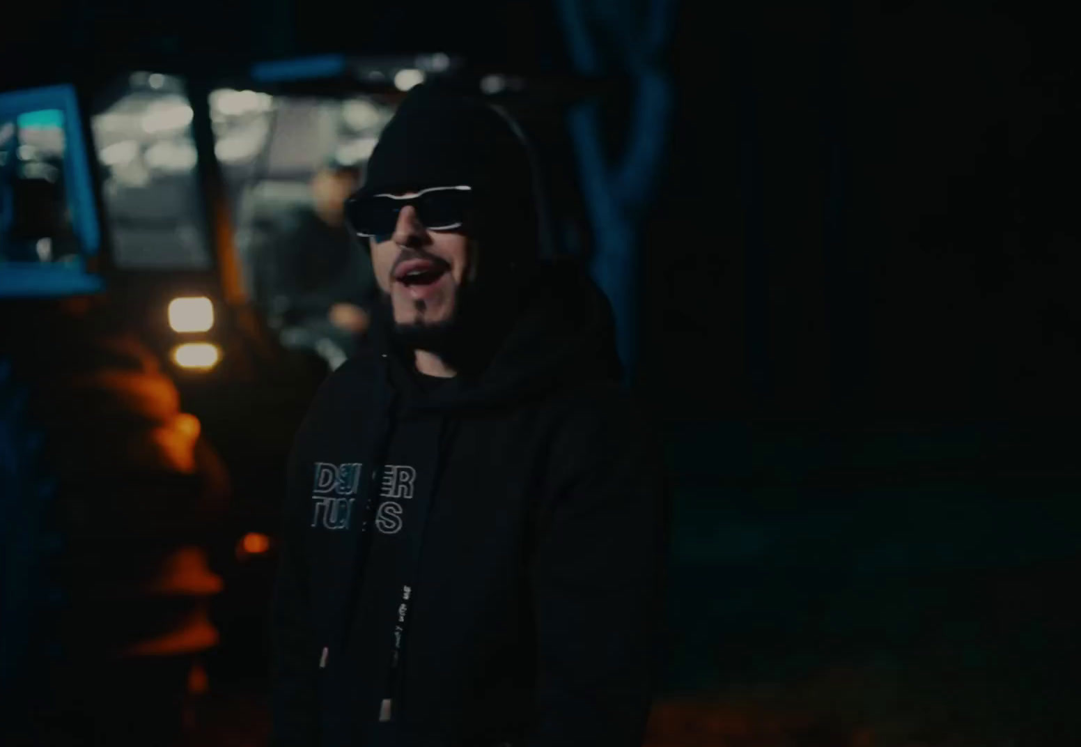 a man wearing sunglasses and a hoodie standing in front of a bus