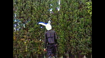 a man in a suit and a white hat standing in front of a hedge