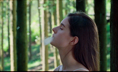 a woman standing in a forest blowing a cigarette