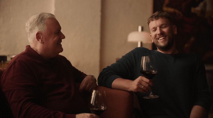 a couple of men sitting next to each other holding wine glasses