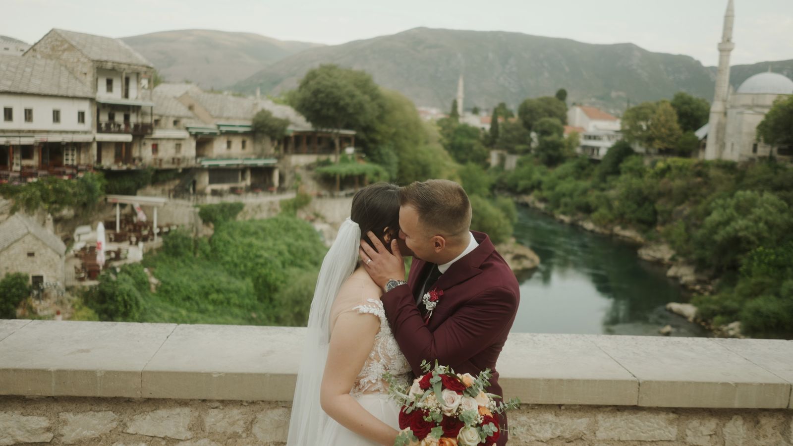 a bride and groom kissing on a bridge overlooking a river