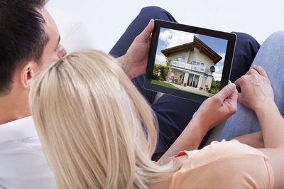 a man and a woman sitting on a couch looking at a house on a tablet