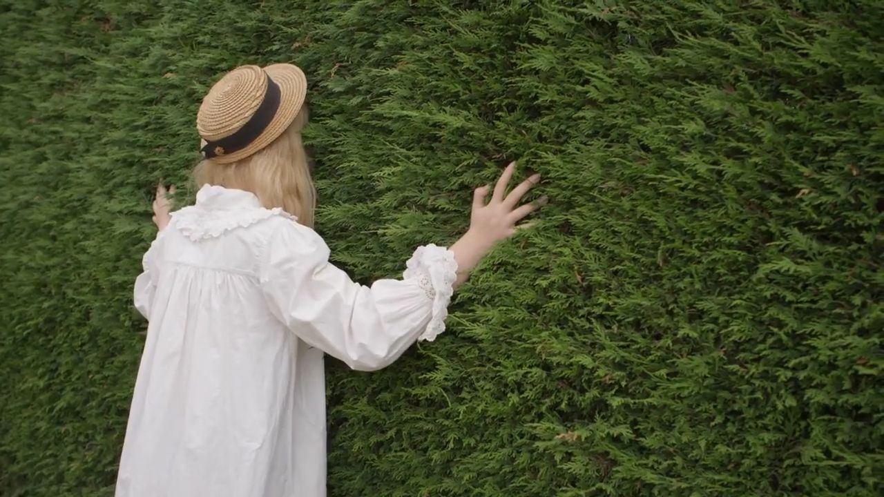 a woman in a white dress and hat standing in front of a hedge