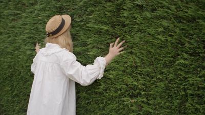 a woman in a white dress and hat standing in front of a hedge