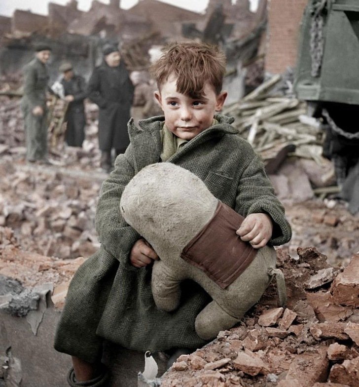 a young boy sitting on a pile of rubble holding a stuffed animal