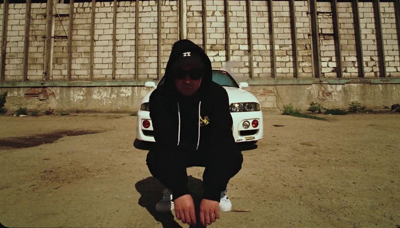 a man in a black hoodie squatting in front of a white car