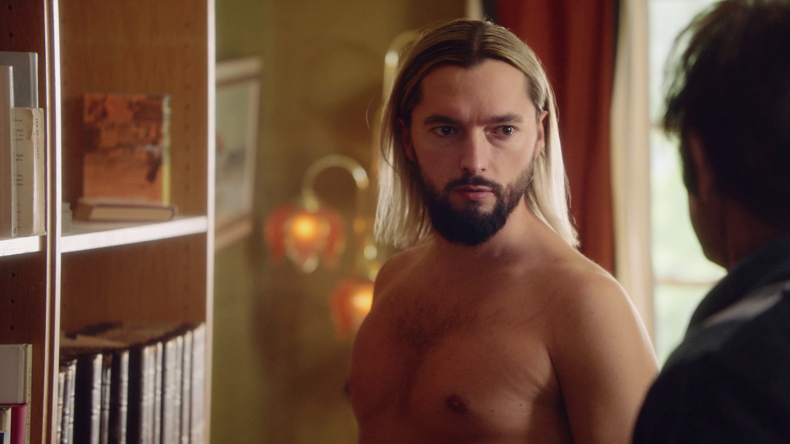 a man with long hair and no shirt standing in front of a bookshelf