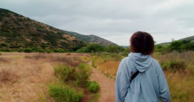 a woman walking down a dirt road next to a lush green hillside