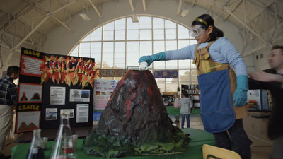 a woman in a blue apron and yellow apron standing next to a fake volcano