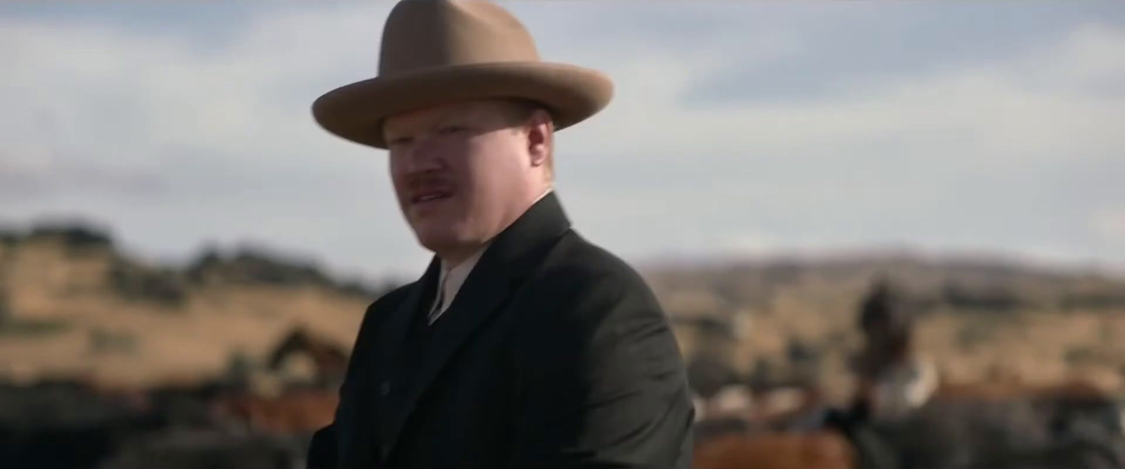 a man in a suit and hat standing in front of a herd of horses