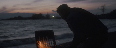 a man standing over a fire next to a body of water